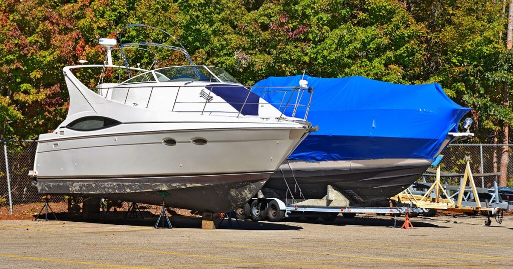 Two boats parked next to each other in a parking lot on a sunny day. One boat is sitting on a trailer and has a blue cover.