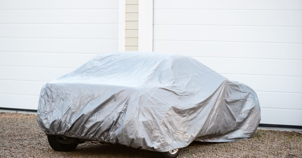 A sedan with a silver protective cover parked on a gravel driveway in front of two closed white garage doors.