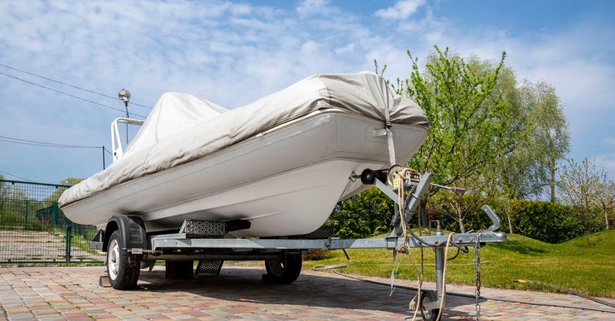 A white boat with a white cover sitting on an elevated metal trailer with wheels on a brick driveway.