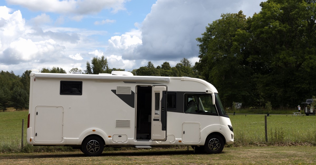 A white and black RV with an open side door parked in front of a grassy field and mature trees on a sunny day.